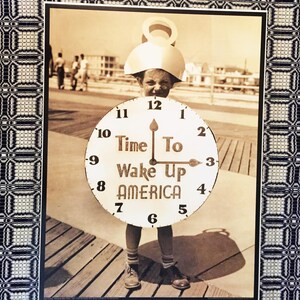 May include: A child dressed as a clock holds a sign that reads "Time To Wake Up America". The child is standing on a wooden boardwalk. The image is in black and white.