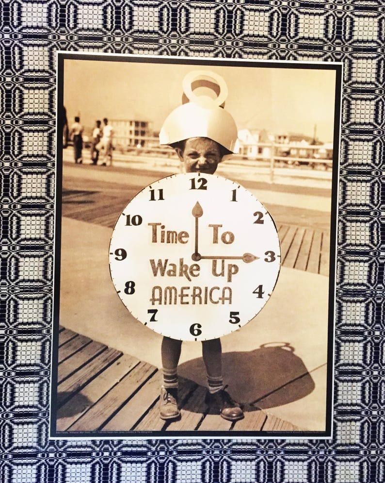 May include: A child dressed as a clock holds a sign that reads "Time To Wake Up America". The child is standing on a wooden boardwalk. The image is in black and white.