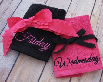 May include: Two embroidered hand towels, one black with pink embroidery that says "Friday" and one pink with black embroidery that says "Wednesday". The towels are tied together with a pink ribbon.