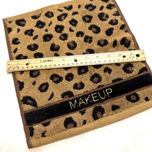 May include: A brown and black leopard print hand towel with a black band and gold embroidery that says "MAKEUP".