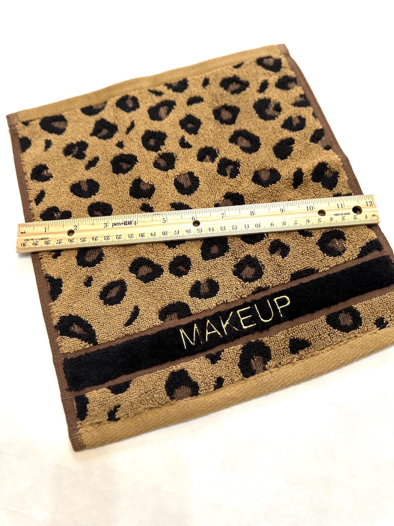 May include: A brown and black leopard print hand towel with a black band and gold embroidery that says "MAKEUP".