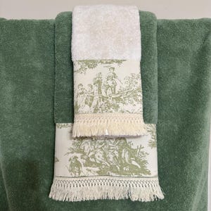 Puede incluir: Un juego de dos toallas verdes y blancas con un patrón de toile. Las toallas tienen un ribete de flecos y están hechas de un material suave y absorbente.