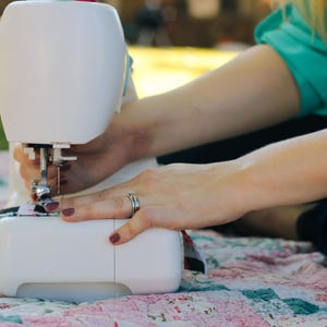 May include: A white sewing machine with a foot pedal is being used to sew a piece of fabric. The machine is sitting on a pink and white patchwork quilt.