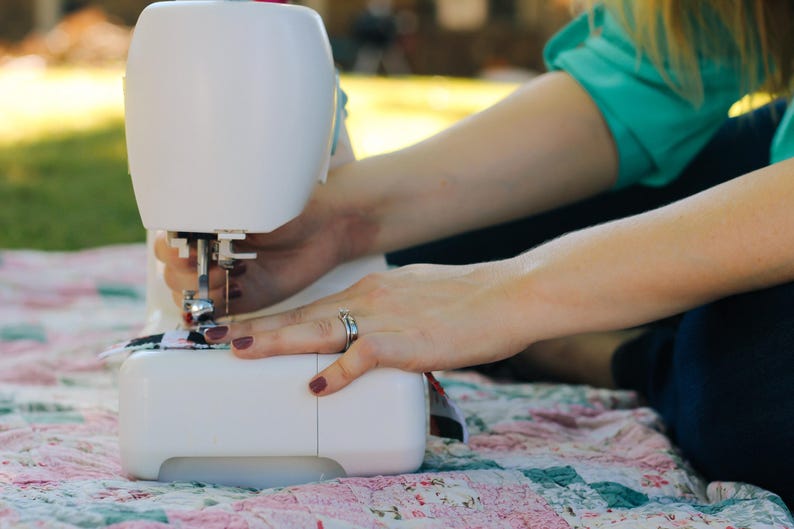 May include: A white sewing machine with a foot pedal is being used to sew a piece of fabric. The machine is sitting on a pink and white patchwork quilt.