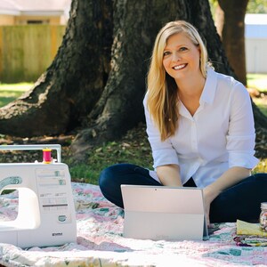 May include: A woman sits on a blanket in a grassy area, using a laptop computer. A sewing machine and a jar of colourful buttons are on the blanket.