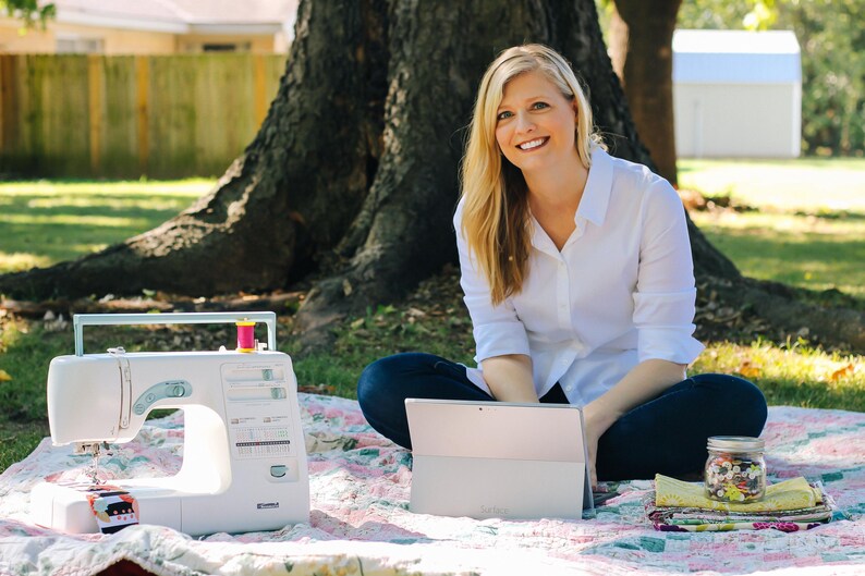 May include: A woman sits on a blanket in a grassy area, using a laptop computer. A sewing machine and a jar of colourful buttons are on the blanket.