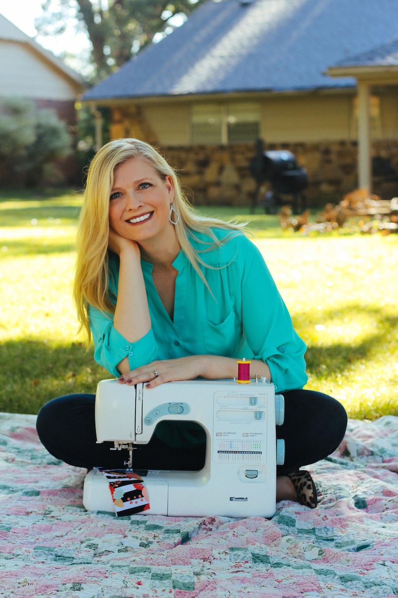 May include: A woman sits on a patterned quilt with a white sewing machine in front of her. The sewing machine has a spool of pink thread on top and a piece of fabric is feeding through the machine.
