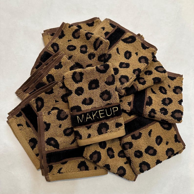 May include: A stack of brown and black leopard print makeup remover cloths with the word "MAKEUP" embroidered in gold on one of the cloths.