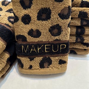 May include: A stack of brown and black leopard print hand towels with the word "MAKEUP" embroidered in gold on the front towel.