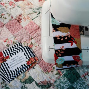 May include: A sewing machine with a black and white fabric featuring red and pink floral accents. The fabric is being sewn onto a white quilt with a patchwork design. The text on the card reads "Augment this towels give your kitchen or bathroom an instant makeover".