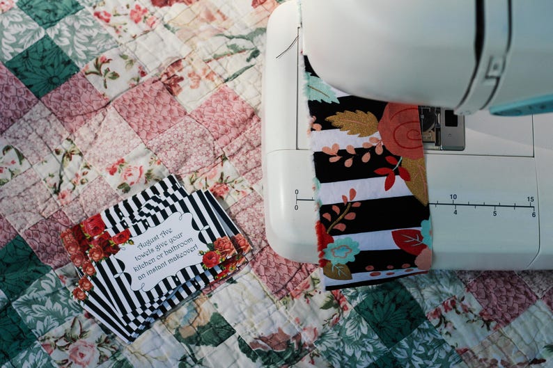 May include: A sewing machine with a black and white fabric featuring red and pink floral accents. The fabric is being sewn onto a white quilt with a patchwork design. The text on the card reads "Augment this towels give your kitchen or bathroom an instant makeover".