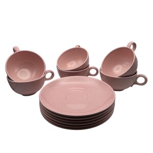 May include: A set of dusty pink ceramic tea cups and matching saucers. The cups feature rounded bowls with small side handles, while the saucers are stacked in a neat pile. The finish on the ceramic pieces is smooth with a subtle speckled texture.