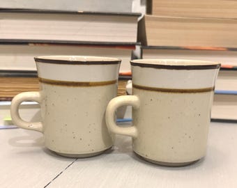 May include: Two vintage ceramic mugs with a speckled beige finish. Each mug has a white rim with a brown band and a matching brown trim. The mugs have a curved handle and are set against a backdrop of stacked books.