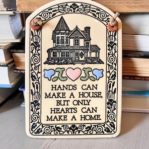 May include: Cream-colored ceramic plaque with a house illustration and the text "HANDS CAN MAKE A HOUSE, BUT ONLY HEARTS CAN MAKE A HOME." The plaque has a decorative border and a jute string for hanging.