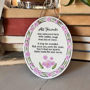 May include: Oval ceramic wall plaque with the text "All Friends" and a poem about welcoming friends. The plaque is white with a border of pink flowers and green leaves. A brown string is attached for hanging.