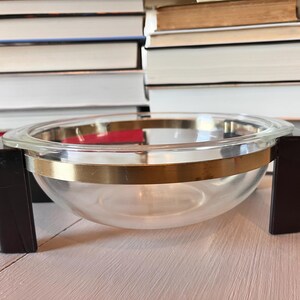 May include: A clear glass serving bowl with a gold-colored band around the middle and dark brown handles. The bowl is sitting on a light-colored surface, with stacks of books in the background.
