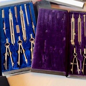 May include: A collection of vintage drafting tools, including compasses and dividers, arranged in velvet-lined cases. The cases are in shades of blue and purple, with the tools appearing to be made of brass or a similar metal.