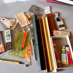 May include: A collection of vintage drafting tools, including rulers, triangles, stencils, and drawing leads. The image features boxes of drawing leads, a yellow TEC drawing lead pencil, and tubes of paint. The tools are arranged on a light gray surface.