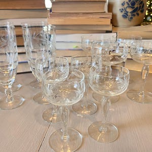 May include: A collection of clear, etched glassware, including champagne coupes, wine glasses, and smaller stemmed glasses. The glasses feature a delicate floral design and are arranged on a light-colored surface.