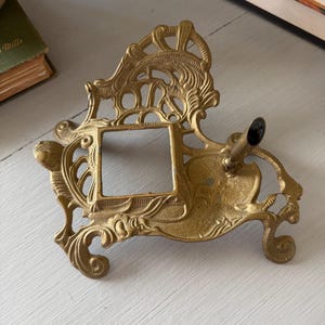 May include: A gold-colored metal desk accessory featuring an ornate, openwork frame with a central square opening and a single cylindrical pen holder attached to the side. The piece has a textured surface and rests on four decorative, curved feet.