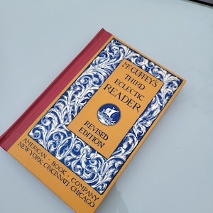 May include: A vintage, yellow and blue book titled "McGuffey's Third Eclectic Reader, Revised Edition" with a decorative border and the text "American Book Company, New York, Cincinnati, Chicago".