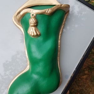 May include: A green and gold Christmas stocking with a tassel. The stocking is made of ceramic and has a gold trim. It is a vintage style stocking.