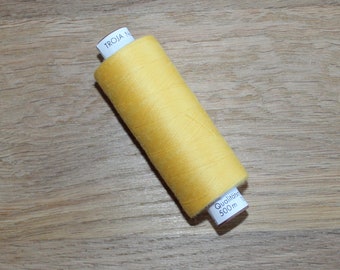 Könnte beinhalten: Eine Spule leuchtend gelben Fadens, beschriftet mit "TROJA N" und "Qualität 500m". Der Faden ist eng auf eine weiße Spule gewickelt, die auf einer hellbraunen Holzoberfläche liegt. Der Faden hat eine lebendige, einfarbige Farbe.