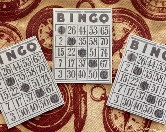 May include: Three vintage bingo cards with the numbers 4, 26, 44, 53, 65, 2, 20, 35, 58, 70, 15, 22, FREE, 51, 74, 7, 17, 37, 56, 67, 3, 30, 40, 50, 62 printed on them. The cards are on a fabric background with a compass design.
