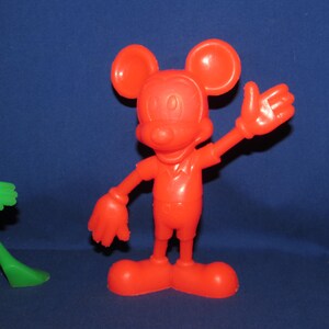 May include: A red plastic toy figurine of Mickey Mouse with large ears and a friendly smile. The toy is standing with one arm raised and a hand outstretched.