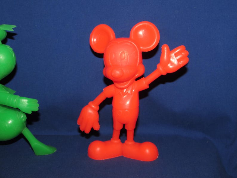 May include: A red plastic toy figurine of Mickey Mouse with large ears and a friendly smile. The toy is standing with one arm raised and a hand outstretched.