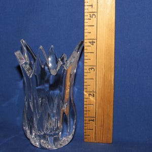 May include: Clear glass vase with a unique, textured design and a jagged, starburst-like rim. The vase is approximately 4 inches tall, as measured by a wooden ruler in the image. The vase is set against a blue background.