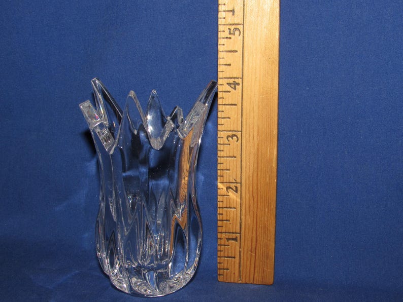 May include: Clear glass vase with a unique, textured design and a jagged, starburst-like rim. The vase is approximately 4 inches tall, as measured by a wooden ruler in the image. The vase is set against a blue background.