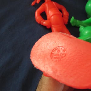 May include: Close-up of the bottom of a red plastic toy figure. The figure has a circular imprint that reads "PLAY-DOH" and "MADE IN USA".