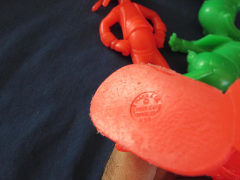 May include: Close-up of the bottom of a red plastic toy figure. The figure has a circular imprint that reads "PLAY-DOH" and "MADE IN USA".