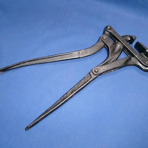 May include: A black metal fencing tool featuring two long handles and a complex hinged head mechanism. The tool has a textured, dark finish and includes visible rivets and pivot points that allow the jaws to open and close for wire tensioning.