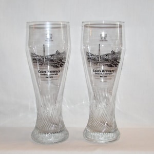 May include: Two clear, tall beer glasses with a twisted base design. Each glass features a black and white graphic of the Coors Brewery in Golden, Colorado, with the brewery's name and establishment date printed below.