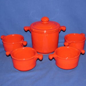 May include: A bright orange ceramic set consisting of one lidded pot and four matching bowls. Each piece features two small, rounded side handles. The pot has a circular lid with a central knob, and all items have a smooth, glossy finish.
