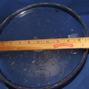 May include: A clear glass cake plate with a scalloped edge and a decorative pattern of floral designs and musical notes. The plate is 9 inches in diameter and is made by Hugh M. Woods.