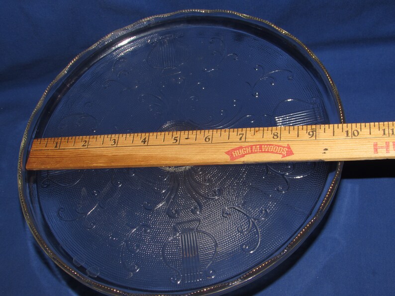 May include: A clear glass cake plate with a scalloped edge and a decorative pattern of floral designs and musical notes. The plate is 9 inches in diameter and is made by Hugh M. Woods.