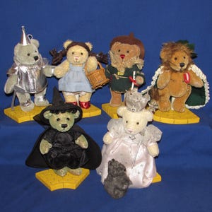May include: A collection of plush teddy bears dressed as characters from The Wizard of Oz. The bears are on yellow brick road bases and include the Tin Man, Dorothy, the Scarecrow, the Cowardly Lion, the Wicked Witch, and Glinda.
