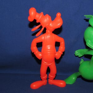 May include: A red plastic toy figure of Goofy standing with his arms crossed. A green plastic toy figure of Donald Duck is standing to the right of Goofy.