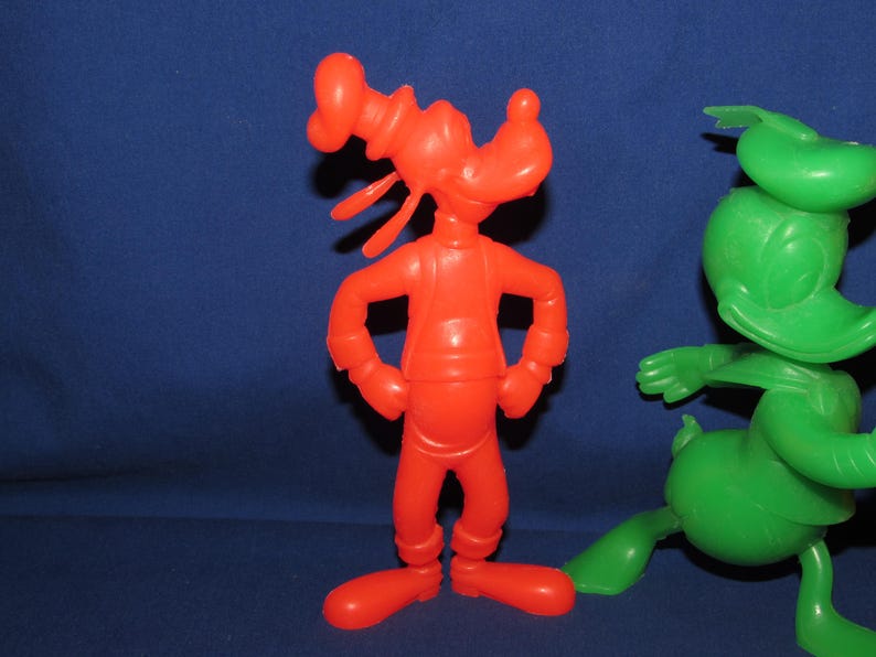 May include: A red plastic toy figure of Goofy standing with his arms crossed. A green plastic toy figure of Donald Duck is standing to the right of Goofy.