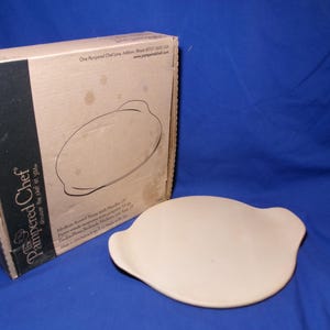 May include: A round, light beige stoneware baking stone with two integrated side handles, accompanied by its original cardboard packaging box. The box features the Pampered Chef logo and text, identifying the item as a 15-inch round stone.