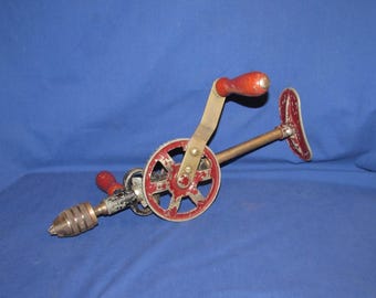 Puede incluir: Un taladro manual antiguo con un mango de madera rojo y una rueda roja y plateada. El taladro tiene un marco de metal y un portabrocas para sujetar las brocas. La herramienta está diseñada para el funcionamiento manual, probablemente para carpintería o proyectos de bricolaje.
