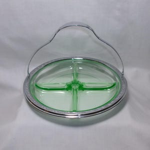 May include: A round, light green glass divided serving dish with four sections. The dish is set within a polished silver-tone metal frame that features a decorative embossed border and a tall, arched metal handle for carrying.