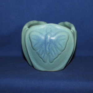 May include: A light blue ceramic planter with a butterfly design. The planter has a rounded shape with a butterfly relief on the front. The color is a muted, seafoam green-blue. The planter is likely for small plants or flowers.