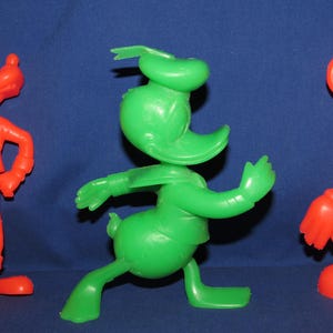 May include: Three vintage plastic cartoon character figurines. A red Goofy, a green Donald Duck, and a red Mickey Mouse.