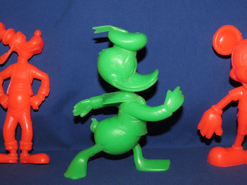 May include: Three vintage plastic cartoon character figurines. A red Goofy, a green Donald Duck, and a red Mickey Mouse.