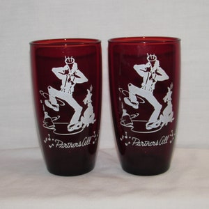 May include: Two ruby red drinking glasses with a white graphic of a dancing figure and a dog. The text "Partners All" is below the graphic. The glasses are cylindrical and appear to be vintage.