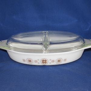 May include: White oval casserole dish with a clear glass lid. The dish has brown floral designs and handles. The lid is divided into two sections. The dish is on a blue background.